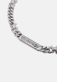 Gros plan d'un bracelet chaîne en argent avec une plaque rectangulaire gravée "ED HARDY" en police stylisée sur un fond uni.