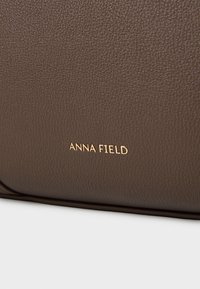 Brown textured faux leather bag with gold "ANNA FIELD" branding on the front. Smooth finish and structured design, minimalistic style.