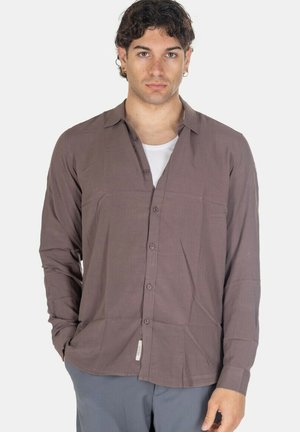 Giovane uomo con capelli ricci che indossa una camicia viola con bottoni sopra una maglietta bianca, in piedi con una mano in tasca davanti a uno sfondo bianco.