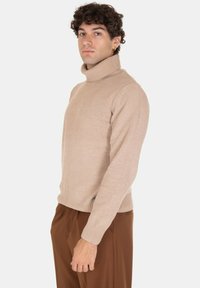 Maglione a collo alto beige con texture a coste e design aderente. Il modello lo indossa con pantaloni marroni, evidenziando una silhouette semplice e pulita.