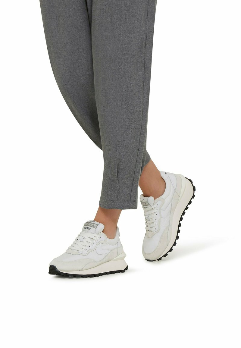 Weiße Sneakers aus einer Mischung von Leder und Wildleder, mit einer strukturierten Sohle und einer gepolsterten Zunge. Kombiniert mit grauen, eng zulaufenden Hosen.