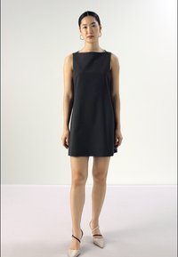 Čierne bezrukávové šaty so štvorcovým výstrihom a rovnými líniami. Hladká textúra, minimalistický dizajn. Modelka má na sebe svetlé špicaté lodičky.