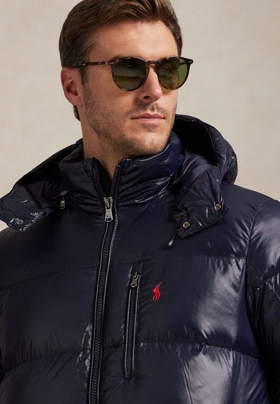 Man wearing dark blue puffer jacket with hood and green-tinted sunglasses, looking slightly to the side.