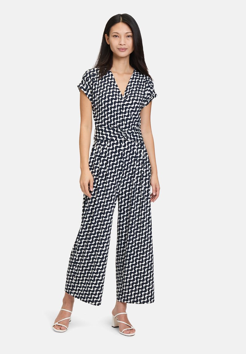 Marinblå jumpsuit med vitt vågigt mönster, korta ärmar, V-ringning och vida ben. Modellen bär vita sandaletter med remmar.