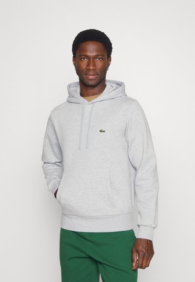 Sweat à capuche gris avec cordons, poche kangourou et un petit logo vert sur la poitrine. Tissu doux et texturé. Associé à un pantalon vert.