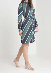 Meikkimekko vihreissä, tummansinissä ja oransseissa raidoissa. Ominaisuuksina pitkät hihat, kaulus ja yhteensopiva vyö. Tyylitetty hihnatoneilla beigen värisillä sandaaleilla.