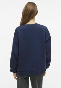 Mornarsko plava oversized dukserica s okruglim izrezom, spuštenim ramenima i rebrastim manžetama. Glatka tekstura, jednostavan dizajn.