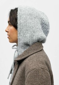 Gorro de punto gris claro con un acabado texturizado y cordón, combinado con un abrigo de lana marrón que presenta un cuello con muescas y detalles en la costura del hombro.