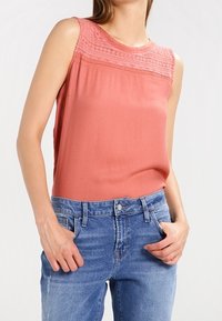 Ärmlös, korallfärgad topp med texturerad spetspanel vid axlarna, matchad med ljusblå denimjeans; mjukt tyg och avslappnad passform.