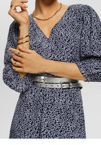 Námořnické šaty s bílým vlnkovaným vzorem, nafouknuté rukávy a stříbrný metalický opasek. Doplňky zahrnují zlaté a stříbrné prsteny a zlatý řetízek.