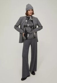 Grau maßgeschneiderte Blazer über einem schwarz-grau gemusterten Strickpullover, kombiniert mit weit geschnittenen grauen Hosen und schwarzen Stiefeletten. Graue Strickmütze.