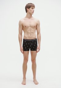 Svarte boksertruser med Polo Ralph Lauren-logo og små hestemønstre. Modellen står barfot foran en nøytral bakgrunn.