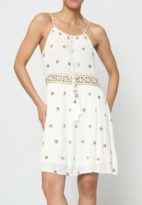 Robe blanche à fines bretelles, ornée de motifs de points dorés métalliques, avec un cordon tressé et un accent à frange à la taille.