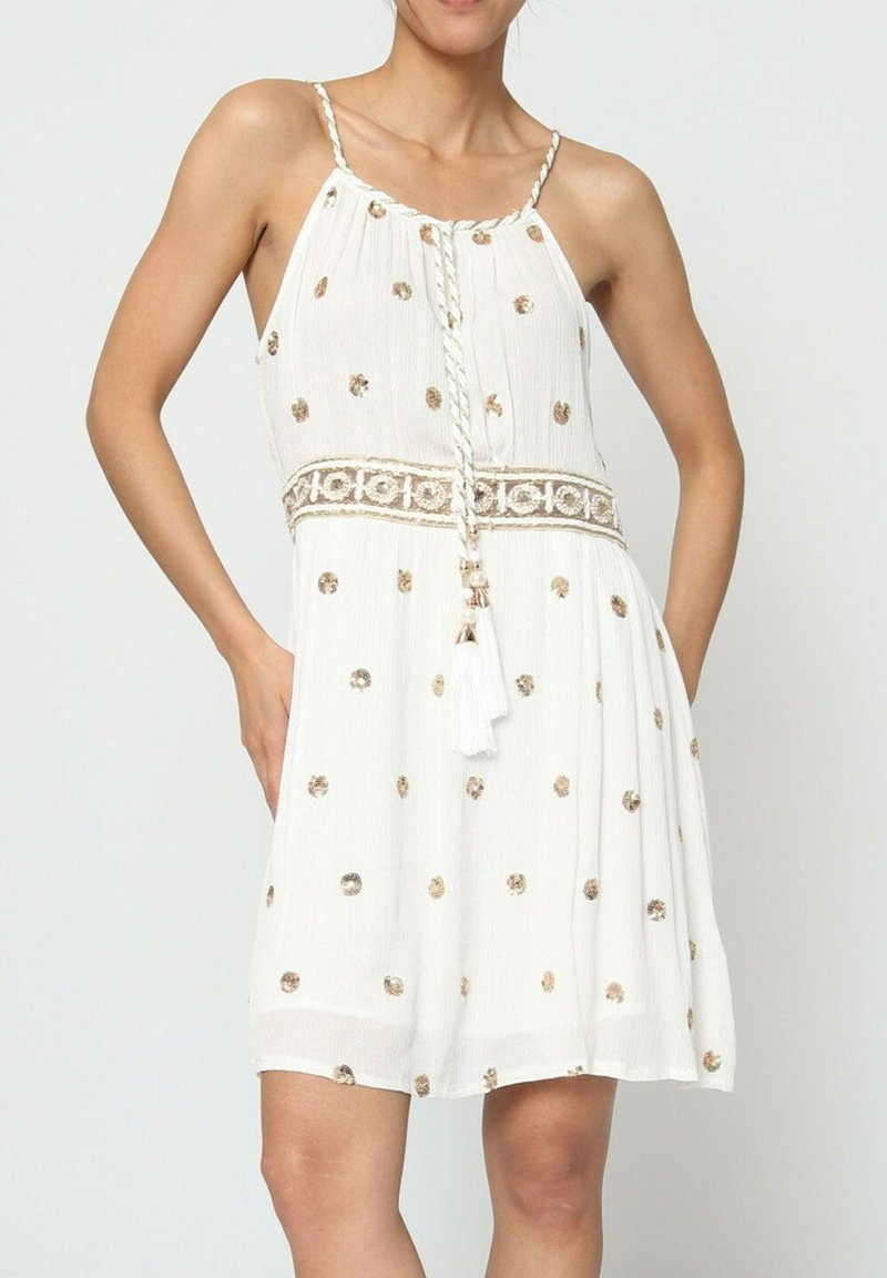 Robe blanche à fines bretelles, ornée de motifs de points dorés métalliques, avec un cordon tressé et un accent à frange à la taille.