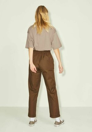 Vrouw met lang blond haar, die een losse taupekleurige top draagt, bruine elastische broek, witte sokken en beige sneakers, staand met de rug naar een lichte muur.