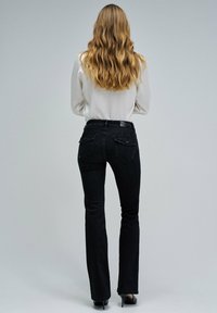 Mujer con cabello rubio largo y ondulado, vestida con una blusa blanca, jeans negros de tiro alto y zapatos de tacón negros, de pie contra un fondo sencillo.