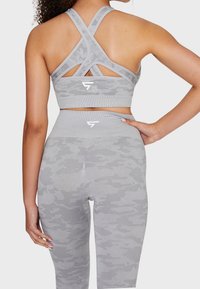 Gray camo sports bra with crisscross straps and logo; high-waisted gray camo leggings with textured pattern and logo detail on waistband.