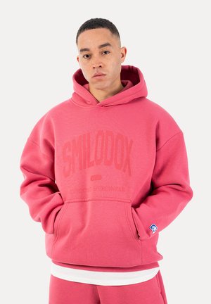 Ung mand med kort hår iført en lyserød hættetrøje og matchende bukser, hænderne i forlommen, neutral udtryk.