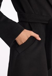 Ręka częściowo włożona w kieszeń czarnej, teksturowanej kurtki z pasującym paskiem, pokazana na białym tle.