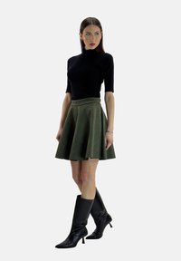 Schwarzes Kurzarmoberteil mit hohem Kragen, kombiniert mit einem grünen Ausgestellt Rock. Das Model trägt glänzende, schwarze Stiefel bis zum Knie. Insgesamt glatte Textur.