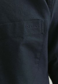 Dunkelblaues Stoffhemd mit strukturierter Oberfläche. Verfügt über eine unauffällige Tasche mit geprägtem "R.D.D."-Logo. Saubere Nähte sind an den Rändern sichtbar.