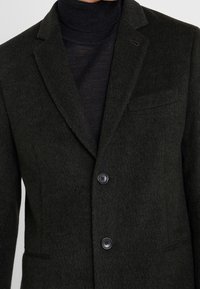 Blazer de lana verde oscuro con un acabado texturizado, dos botones en la parte frontal y un bolsillo en el pecho. Diseño entallado con solapas de pico.