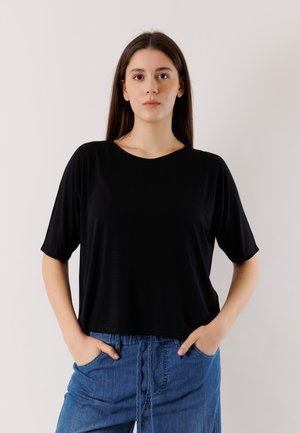 Giovane donna con capelli castani lunghi, che indossa una maglietta nera a maniche corte e pantaloni di jeans blu, in piedi con le mani nelle tasche.