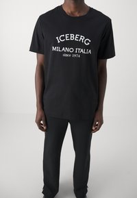 Musta puuvillapaita, jossa on valkoiset tekstit "ICEBERG MILANO ITALIA vuodesta 1974", lyhyet hihat ja pyöreä kaula-aukko.