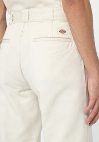 Beige Hose mit aufgesetzten Gesäßtaschen und einem kleinen rechteckigen Dickies-Logo-Patch neben der rechten Tasche, getragen von einer Person mit sichtbarem rechten Hand.