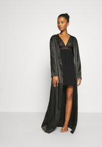 Svart satinrok med långa ärmar över en svart klänning med spetskant. Klänningen har en V-ringning och en flödande kjol. Barfotaställning.