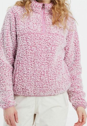 Osoba w jasnoróżowej, teksturowanej polarowej bluzie na zamek z długimi rękawami i białych spodniach, na jednolitym białym tle.