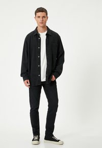 Czarna koszula dżinsowa z zapinaniem na guziki i kieszonką na piersi, noszona na białej koszulce. Czarne jeansy slim fit i czarne trampki z białą podeszwą.