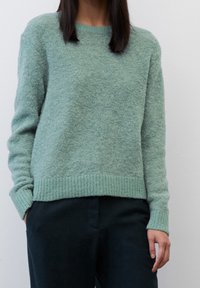 Eine Frau trägt einen strukturierten, hellgrünen Pullover und dunkle Hosen, während sie mit den Händen in den Taschen vor einem schlichten, hellen Hintergrund steht.