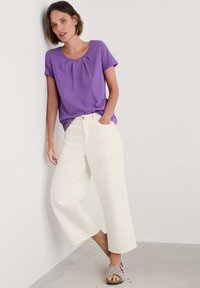 Top morado de manga corta con escote en forma de llave, combinado con jeans blancos de pierna ancha y holgados. La modelo lleva sandalias grises y está de pie frente a una pared.