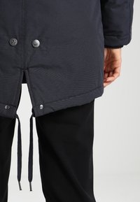 Granatowa odzież wierzchnia o teksturowanej powierzchni, z zatrzaskami i regulowanymi troczkami u dołu. Pod spodem widoczne czarne spodnie.