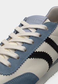 Joggesko med en blanding av blått og kremfarget skinn, svarte detaljer, bølgete design og hvite lisser med teksturerte overflater.