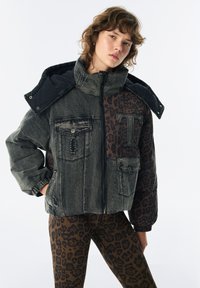 Jeune personne portant une veste à col haut bicolore avec des panneaux en denim usé et imprimé léopard, accompagnée de leggings assortis à imprimé léopard.