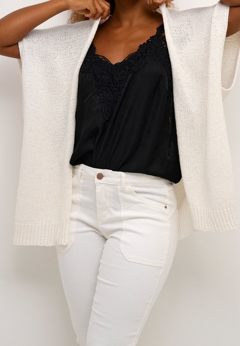 Cárdigan blanco de punto con mangas cortas, llevado abierto sobre una blusa negra con encaje, combinado con pantalones blancos ajustados.