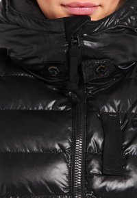 Chaqueta acolchada negra con cuello alto, cremallera metálica y botones. Material suave y brillante con patrones acolchados y correas ajustables.