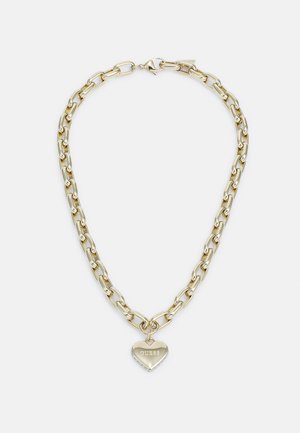 Collier chaîne en or avec de gros maillons et un pendentif en forme de cœur gravé avec le mot "GUESS" sur fond blanc.