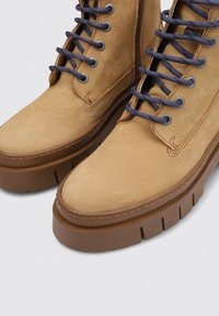 Botas de cuero color marrón claro con cordones oscuros azules y suelas gruesas de goma marrón, vistas desde un ángulo cercano desde arriba.