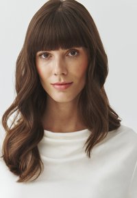 Modelka s dlouhými, vlnitými hnědými vlasy a ofinou, oblečená v bílém topu z drapované látky. Jemná pleť, neutrální make-up a neutrální pozadí.