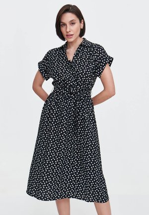 Czarna sukienka w białe kropki, z krótkimi rękawami, kołnierzykiem i przewiązywanym w pasie paskiem. Lekka tkanina o długości midi.