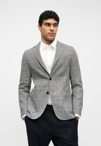 Άνδρας που φοράει γκρι σακάκι με καρό, λευκό πουκάμισο με κουμπιά και μαύρο παντελόνι, στέκεται με τα χέρια στις τσέπες μπροστά σε ένα απλό φόντο.