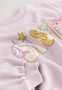 Violets džemperis ar mīkstu struktūru, uz kura ir rotaļīgi grafiski attēli ar vienradzi, zaķi un krāsainām zvaigznēm ar svītrām. Rievots kakla izgriezums un aproces.