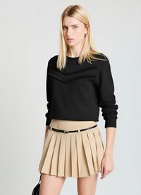 Pull noir avec un motif chevron texturé sur la poitrine, associé à une jupe plissée beige et une ceinture noire. Tissu lisse et coupe ajustée.