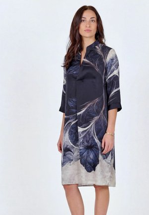 Mujer de pie con un vestido de manga larga hasta la rodilla, con un estampado floral oscuro y un pequeño cuello mandarín, sobre un fondo liso.