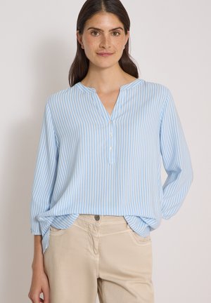 Femme aux cheveux foncés portant un chemisier à manches longues rayé bleu clair et blanc et un pantalon beige sur un fond uni.