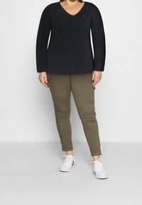 Personne portant un pull noir à manches longues avec col en V, un pantalon cargo olive et des baskets blanches, debout devant un fond uni.