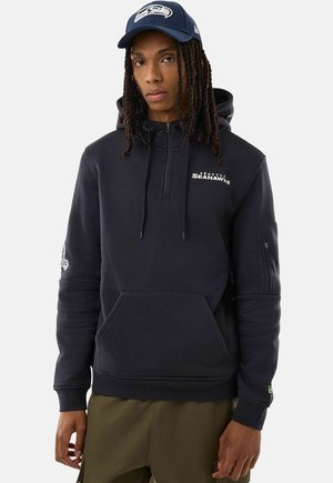 Persona con trecce lunghe indossa un berretto da baseball dei Seattle Seahawks blu navy e una felpa nera con cappuccio con logo sul petto e sulla manica, in piedi su sfondo bianco.